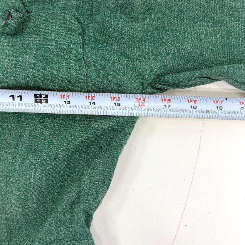 May include: Close-up of a green garment with a measuring tape laid across it. The tape shows measurements in inches, with markings from 11 to 20 inches. The fabric has a textured appearance.