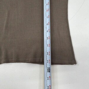 Op de afbeelding: Een effen bruine gebreide top met een fijne ribbelstructuur. Een wit meetlint is verticaal over de stof geplaatst en toont een lengtemeting van 38 cm tot 66 cm, wat de verticale schaal van het kledingstuk aangeeft.