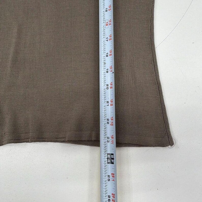 Op de afbeelding: Een effen bruine gebreide top met een fijne ribbelstructuur. Een wit meetlint is verticaal over de stof geplaatst en toont een lengtemeting van 38 cm tot 66 cm, wat de verticale schaal van het kledingstuk aangeeft.