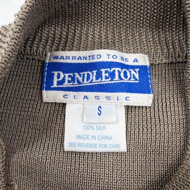 Op de afbeelding: Een bruin gebreid kledingstuk met een wit en blauw Pendleton Classic merklabel. Het label bevat de tekst Warranted to be a Pendleton, maat S, 100% silk, Made in China en See reverse for care. De stof heeft een geribbelde textuur.