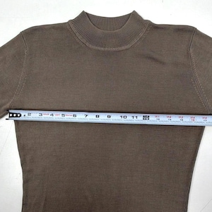 Op de afbeelding: Een bruin shirt met korte mouwen en een opstaande kraag. Een wit meetlint is over de borst geplaatst en toont een breedte van ongeveer 51 cm van oksel tot oksel. Het kledingstuk heeft over het gehele oppervlak een fijne breistructuur.