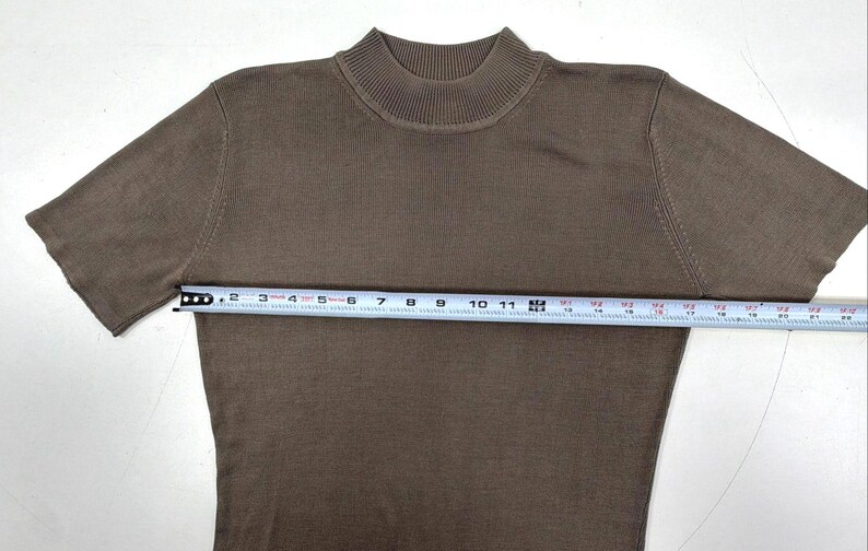 Op de afbeelding: Een bruin shirt met korte mouwen en een opstaande kraag. Een wit meetlint is over de borst geplaatst en toont een breedte van ongeveer 51 cm van oksel tot oksel. Het kledingstuk heeft over het gehele oppervlak een fijne breistructuur.