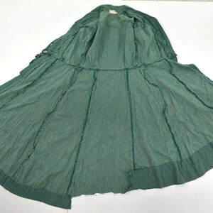 May include: A green, open-front coat with a wide skirt, laid flat on a white surface. The coat has a textured appearance with visible stitching along the seams. The fabric appears to be a lightweight material.
