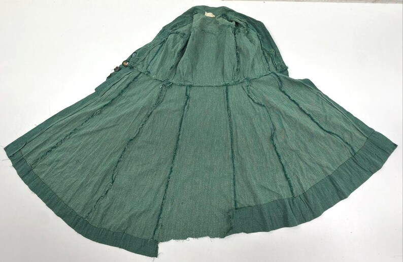 May include: A green, open-front coat with a wide skirt, laid flat on a white surface. The coat has a textured appearance with visible stitching along the seams. The fabric appears to be a lightweight material.