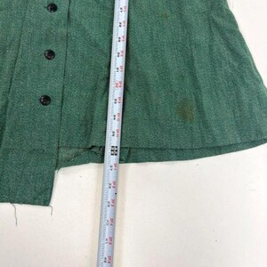 May include: A close-up of a green garment with black buttons and a measuring tape. The garment has a textured appearance and appears to be made of a woven fabric. The measuring tape shows measurements in inches.