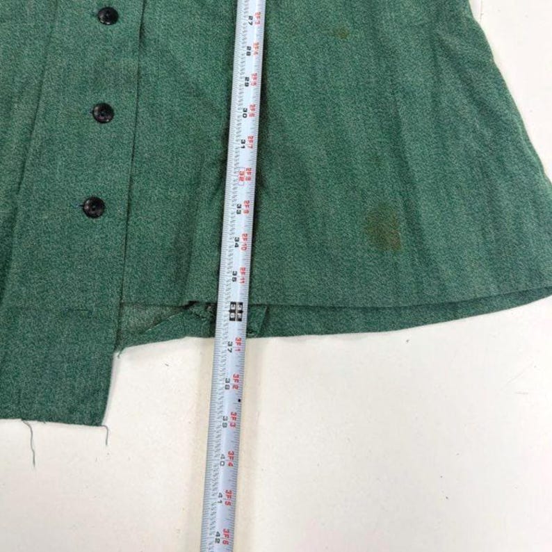 May include: A close-up of a green garment with black buttons and a measuring tape. The garment has a textured appearance and appears to be made of a woven fabric. The measuring tape shows measurements in inches.