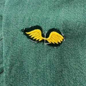 May include: Close-up of a green fabric with a yellow and black embroidered wing patch. The wings are detailed with yellow feathers and outlined in white thread. The fabric has a textured appearance.