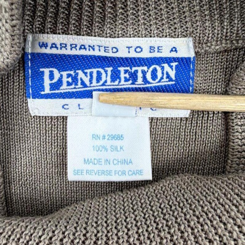 Op de afbeelding: Een bruin gebreid kledingstuk met een wit en blauw Pendleton-merklabel. De tekst luidt WARRANTED TO BE A PENDLETON. Een wit label eronder geeft 100% zijde, RN #29685, MADE IN CHINA en SEE REVERSE FOR CARE aan.