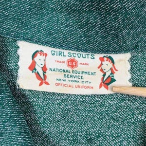 May include: Close-up of a green fabric with a white label. The label reads "Girl Scouts" with illustrations of girls and the text "National Equipment Service, New York City, Official Uniform."