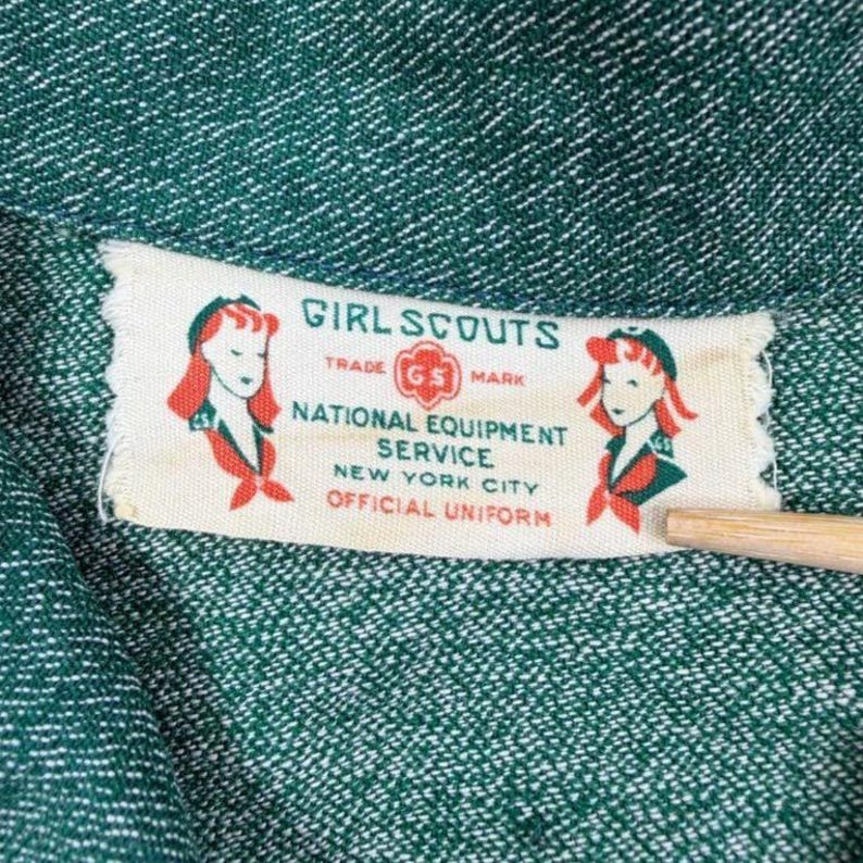 May include: Close-up of a green fabric with a white label. The label reads "Girl Scouts" with illustrations of girls and the text "National Equipment Service, New York City, Official Uniform."