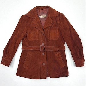 May include: A rust-colored suede jacket featuring a collared neckline, long sleeves with buttoned cuffs, and a matching belt with a rectangular buckle. The front includes two chest pockets, two lower pockets, and a button-down closure with the label the tannery.