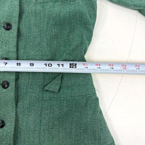 May include: A close-up of a green blazer with black buttons and a pocket. A measuring tape is placed across the blazer, showing measurements in inches.