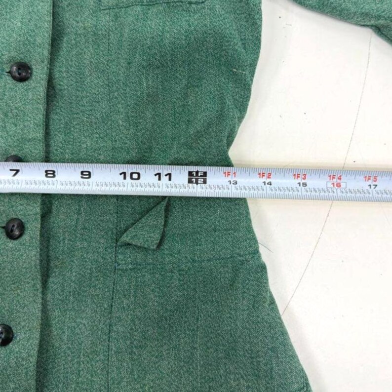 May include: A close-up of a green blazer with black buttons and a pocket. A measuring tape is placed across the blazer, showing measurements in inches.