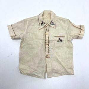May include: A short-sleeved, cream-colored shirt with embroidered details. The shirt features a collar and a pocket with a rabbit design. The collar and pocket are adorned with a brown stitched border. The shirt is buttoned with wooden toggles.