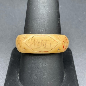 Op de afbeelding: Een lichtbruine, uitgesneden ring met een centraal achthoekig paneel met de gegraveerde letters W.J.H. De band is aan beide zijden versierd met geometrische ruitvormige motieven, geaccentueerd met rood pigment in de lijnen.