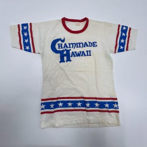 May include: Cream-colored t-shirt with red trim on the collar and sleeves. The sleeves have a blue and red star pattern. The shirt features the text "Chaminade Hawaii" in blue lettering, with a red stripe at the bottom.