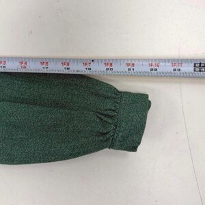 May include: Close-up of a dark green fabric sleeve with a buttoned cuff. A ruler is placed above the sleeve, measuring in inches. The fabric has a textured appearance.