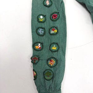 May include: Close-up of a green long-sleeved shirt sleeve adorned with various embroidered patches. The patches feature colorful designs, including flowers, trees, and other nature-themed motifs. The sleeve is made of a textured fabric.