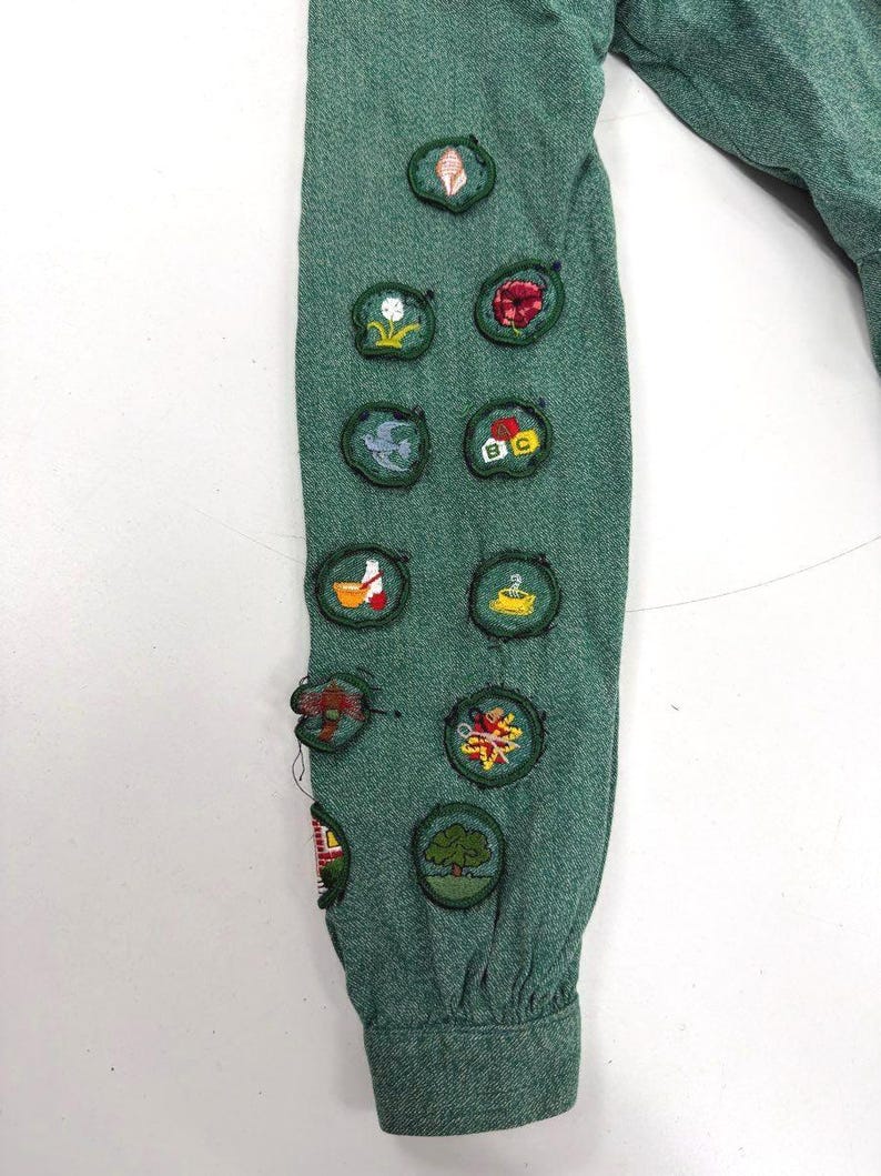 May include: Close-up of a green long-sleeved shirt sleeve adorned with various embroidered patches. The patches feature colorful designs, including flowers, trees, and other nature-themed motifs. The sleeve is made of a textured fabric.