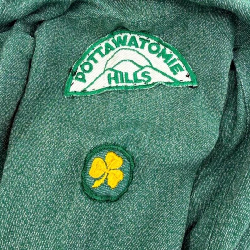 May include: Close-up of a green fabric with two embroidered patches. The top patch is a white and green arched design with the words "POTTAWATOMIE HILLS". The bottom patch is a green circle with a yellow shamrock.