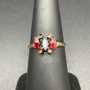 May include: A gold-toned ring featuring a central oval, pale blue stone flanked by two red, marquis-cut stones. Additional small, round accents surround the central stones. The ring is displayed on a black ring holder.