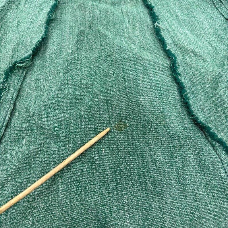 May include: Close-up of green fabric with a rough, frayed edge. A wooden stick points towards a small, dark stain on the textured material. The fabric appears to be a medium weight, possibly denim or a similar woven textile.