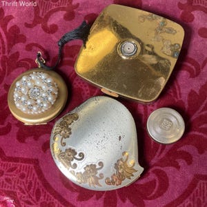 May include: A collection of vintage compacts and a small container on a red patterned surface. The compacts include a gold-toned square compact, a round compact with faux pearls, and a heart-shaped compact with gold floral accents.