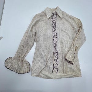 May include: Cream-colored blouse with a small, dark pattern. The long sleeves have ruffled cuffs, and a ruffled trim runs down the front. The collar is pointed, and the blouse has buttoned cuffs.