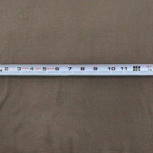 Op de afbeelding: Een effen taupekleurig kledingstuk met een metalen meetlint eroverheen gespannen, wat een breedte van ongeveer 43 centimeter aangeeft van de linkerkant tot de rechterkant van het stoffen oppervlak.