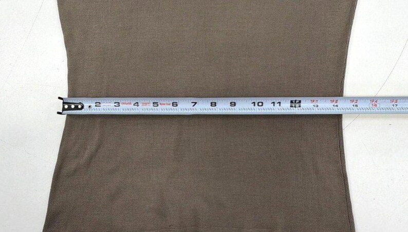 Op de afbeelding: Een effen taupekleurig kledingstuk met een metalen meetlint eroverheen gespannen, wat een breedte van ongeveer 43 centimeter aangeeft van de linkerkant tot de rechterkant van het stoffen oppervlak.