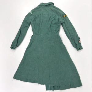 May include: A green long-sleeved dress with a collared neckline and a flared skirt. The dress features patches on the sleeves and a button-down front. The fabric has a textured appearance.