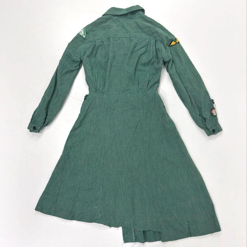 May include: A green long-sleeved dress with a collared neckline and a flared skirt. The dress features patches on the sleeves and a button-down front. The fabric has a textured appearance.