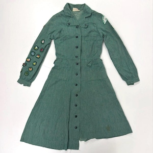 May include: A vintage green dress with long sleeves, a collared neckline, and a button-down front. The dress features a fitted waist, pockets, and a flared skirt. Patches are sewn on the left sleeve. The dress is made of a textured fabric.