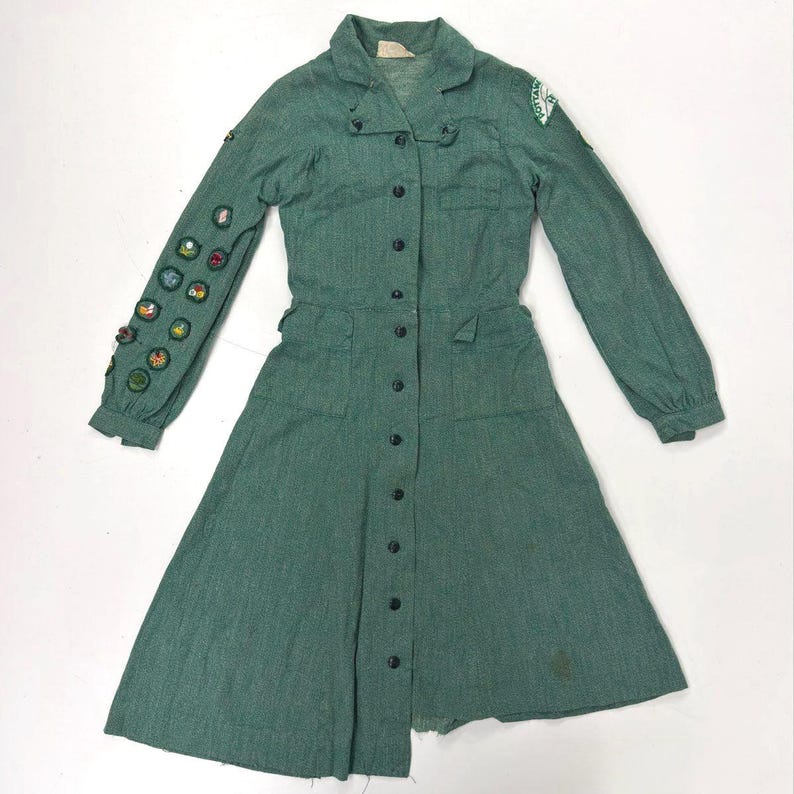 May include: A vintage green dress with long sleeves, a collared neckline, and a button-down front. The dress features a fitted waist, pockets, and a flared skirt. Patches are sewn on the left sleeve. The dress is made of a textured fabric.