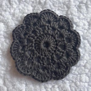 May include: A dark gray, handmade crochet coaster with a floral design. The coaster is round with a scalloped edge and a central circular pattern. The yarn appears to be a soft, textured material.