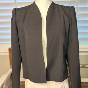 May include: A dark gray blazer with a notched collar and long sleeves. The blazer has shoulder pads and a tailored fit. The jacket is displayed on a mannequin, showcasing its structure and design. The blazer is a classic piece of apparel.