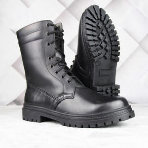 Könnte beinhalten: Ein Paar schwarze Leder-Kampfstiefel. Die Stiefel haben ein Schnürdesign, eine dicke Profilsohle und eine mittlere Wadenhöhe. Ein Stiefel steht aufrecht, der andere ist abgewinkelt, um das Profil der Sohle zu zeigen.