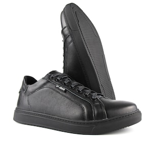 Puede incluir: Zapatillas de cuero negras de corte bajo. Los zapatos tienen una puntera redondeada, cordones negros y una suela gruesa negra. La marca "U-Bot" es visible. Un zapato está en ángulo para mostrar la suela.