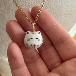 May include: A gold-toned chain necklace featuring a small, white, rounded ceramic cat pendant. The cat has closed eyes, pink cheeks, red ear tips, and floral accents. The Chinese character for wealth, 財, is painted in gold on the cat's forehead.