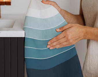 May include: A folded, rectangular towel with a gradient of blue hues, from light to dark, separated by thin white lines. The towel is held up by a person. The towel is likely for use in a bathroom or kitchen.