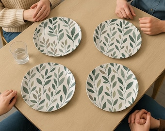 May include: Four white ceramic plates with a green and gray leaf pattern are arranged on a light brown wooden table. The plates are round and have a repeating design of stylized leaves and branches. A clear glass of water sits on the table.