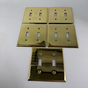 May include: A set of five polished brass double toggle light switch plates. Each rectangular plate features two vertical rectangular cutouts for switches and four small circular holes for mounting screws, arranged in a symmetrical grid pattern.
