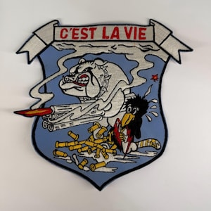 May include: An embroidered patch featuring a light blue shield shape with a banner at the top reading C'EST LA VIE in red. The design depicts a white bulldog firing a large gun with smoke, yellow bullet casings, and a black bird character.
