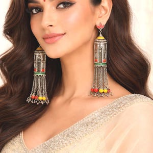 May include: Silver-toned dangle earrings with intricate detailing. The earrings feature a rectangular design with red, green, and yellow accents, and dangling chains with colorful beads. The earrings are worn by a person.