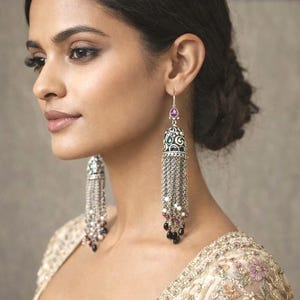 May include: Elaborate silver earrings with long chain tassels and decorative tops. The earrings have a purple gemstone and are embellished with white, black, and red beads. The model wears a cream-coloured, embroidered top with floral patterns.