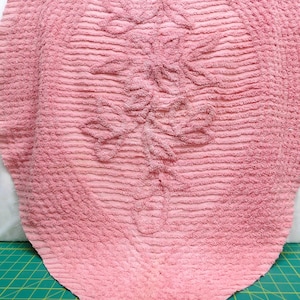 May include: A vintage pink bath mat with a textured, ribbed surface and a floral design in the center. The mat has a rounded top and a pointed bottom. The mat is made of a soft, absorbent material.
