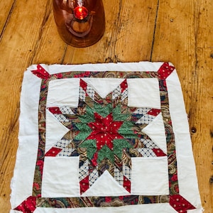 May include: A square fabric quilt block featuring a central eight-pointed star pattern in red, green, and patterned fabrics. The star is surrounded by a white border with a decorative paisley and floral print frame and red corner accents.