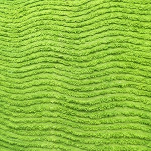 May include: Close-up of a bright green, textured fabric with a wave-like pattern. The fabric has a soft, plush appearance, with visible ridges and a vibrant, solid color. The texture suggests a cozy or decorative textile.