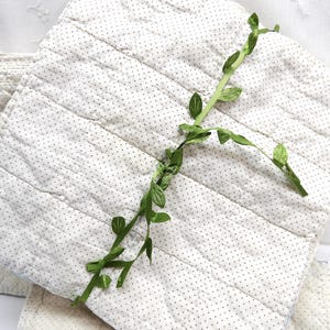 May include: A stack of square, quilted fabric pieces in a light cream color with small, evenly spaced dark gray dots. A green, leafy vine is draped across the top piece. The fabric has a stitched, horizontal pattern.