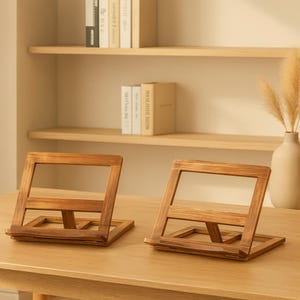 May include: Two wooden book stands with a natural finish are displayed on a light-colored wooden table. The stands are angled, with a rectangular base and a frame to hold a book. The background includes a shelf with books and a vase with dried plants.